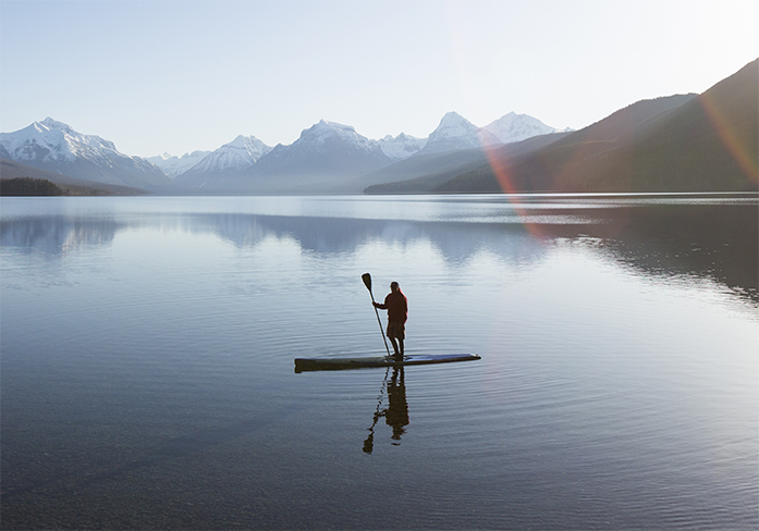 A man standing on a paddle board, in a lake surrounded by mountains.